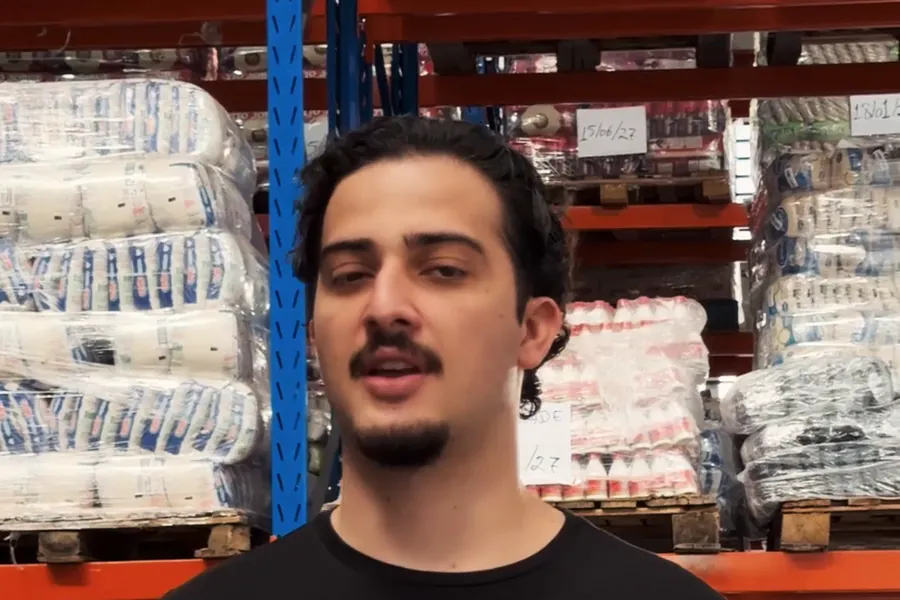 O corredor de um galpão de distribuidora, com prateleiras de paletes cheios de sacarias e fardos, e Gabriel Medeiros no centro.