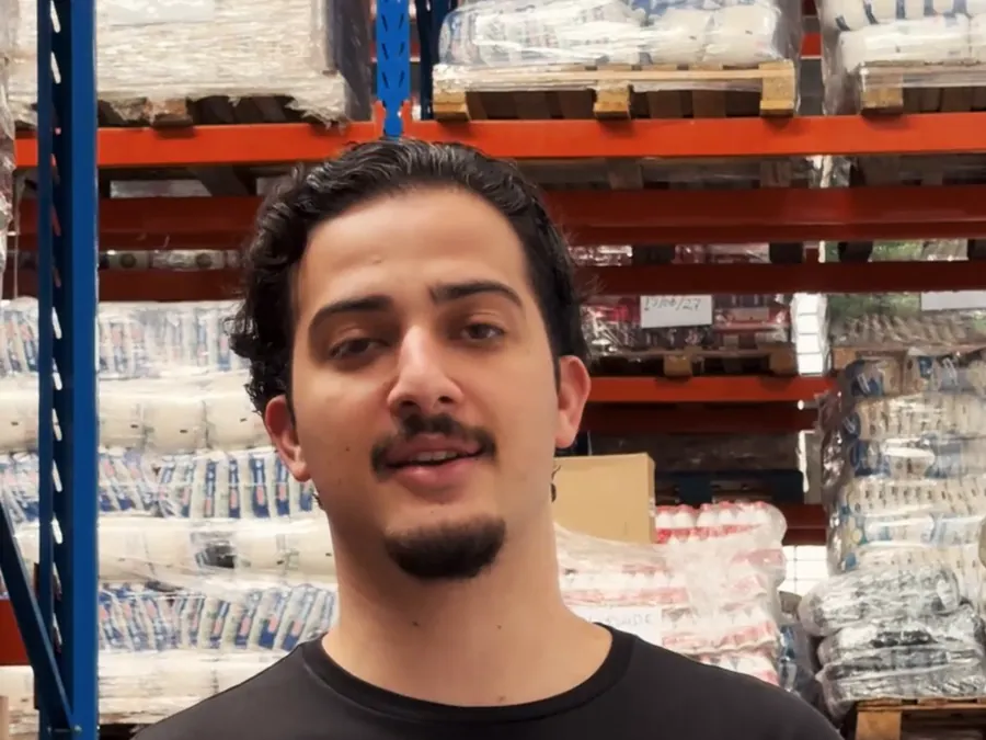 Gabriel Medeiros falando no galpão de uma distribuidora, mais de perto, com paletes de sacarias ao fundo.